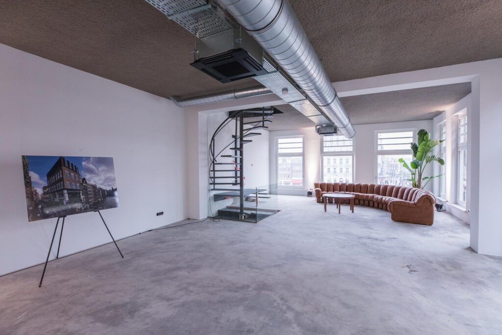 Ruime, industriële loft aan de Prins Hendrikkade met grote ramen, een spiraaltrap, bruine hoekbank en een impressieafbeelding van het gebouw op een ezel.